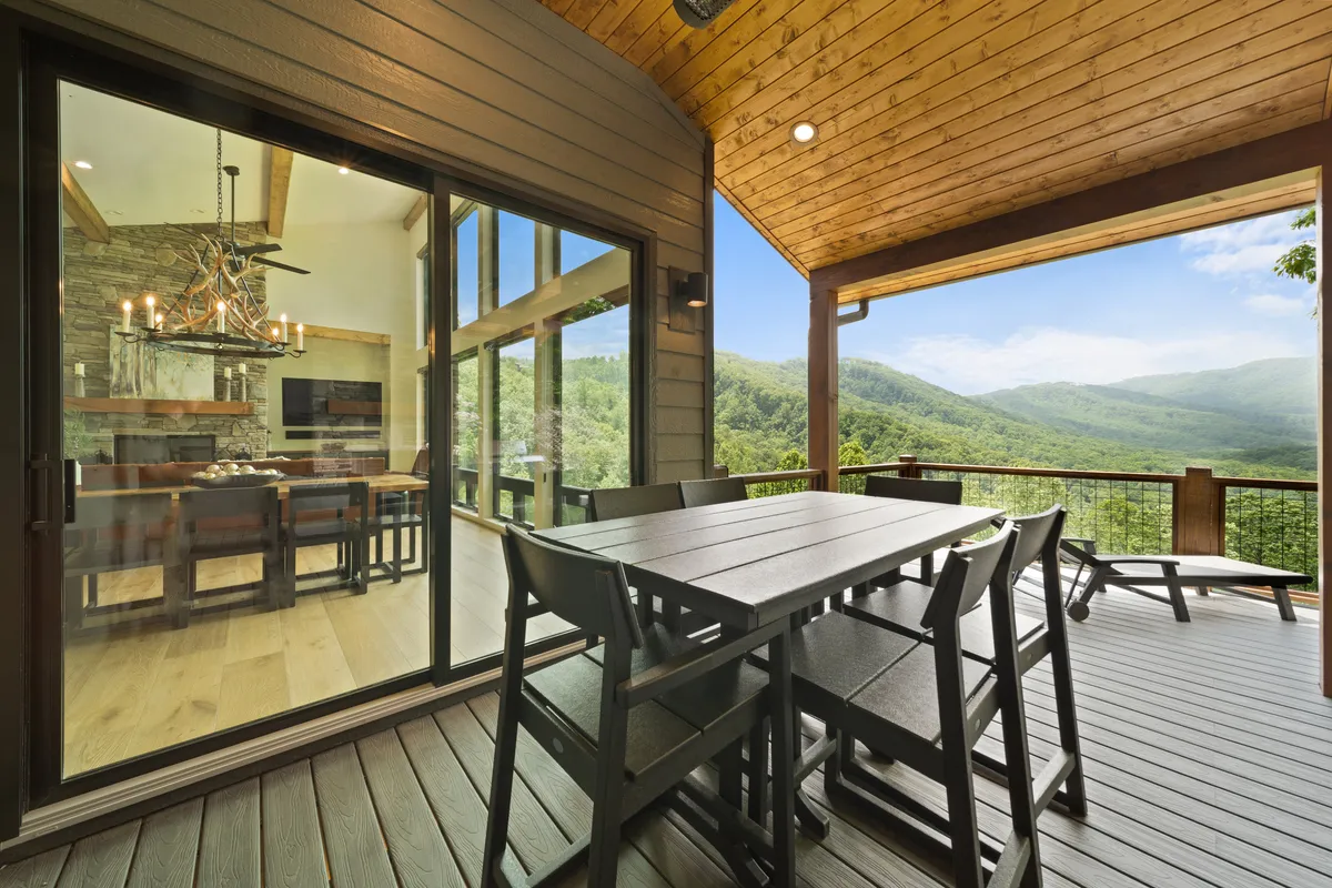An inviting outdoor dining table sits beneath a wooden ceiling, opening to panoramic mountain views. Large glass doors connect the patio to the interior, creating a seamless indoor-outdoor living experience.