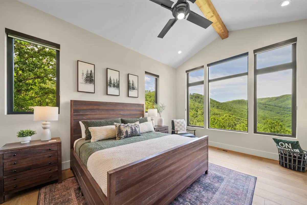 A cozy bedroom with a bed dressed in plush pillows and linens, large windows showcasing mountain views, a nightstand with a lamp, and framed artwork on the walls. The setting is tranquil and filled with natural light.
