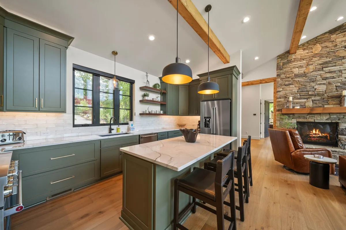 A modern kitchen featuring a spacious island with bar stools, sleek appliances, and attractive stone and wood accents. The kitchen is perfectly equipped for gourmet cooking and casual gatherings alike, with sightlines into the adjacent living spaces and a stone fireplace anchoring the area.