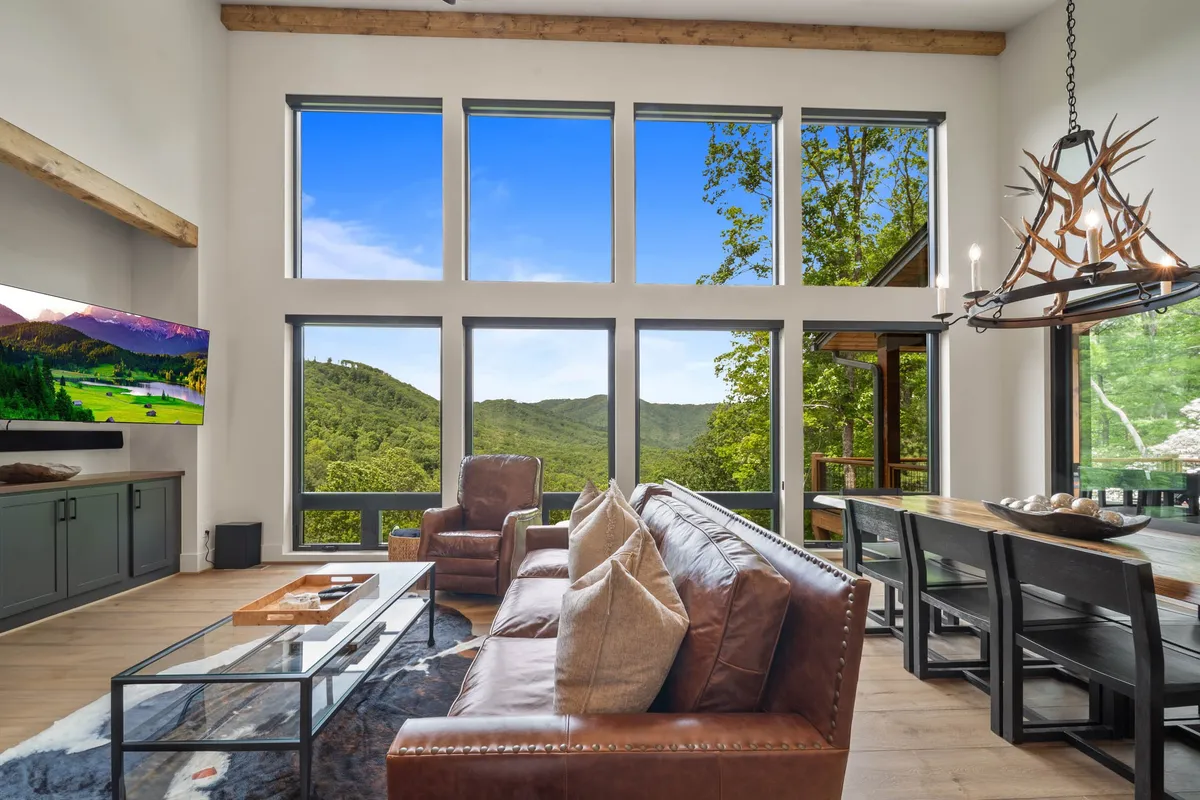 A spacious living and dining area featuring large windows that let in an abundance of natural light. The room is centered around a cozy leather sofa and a dining table beneath a statement chandelier. You can see a television and touches of homey decor, and the windows provide a clear view of the mountains beyond. The space is both elegant and welcoming, with a clear connection to the outdoors.