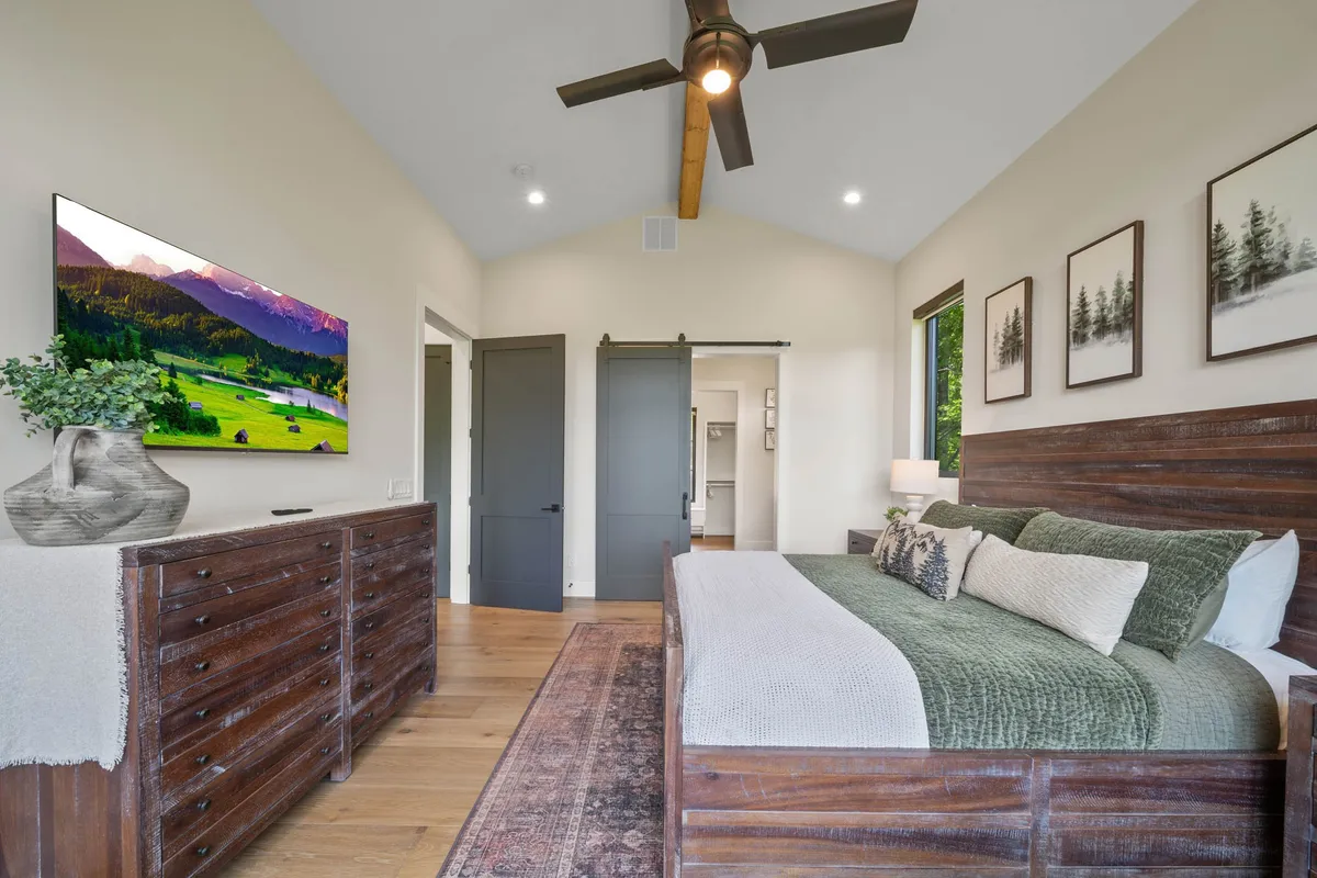 A serene bedroom featuring a wooden bed frame with green accent pillows, a TV displaying a peaceful landscape, dark closet doors, and tasteful artwork on the walls. The room is illuminated by natural light and exudes a tranquil atmosphere perfect for rest.