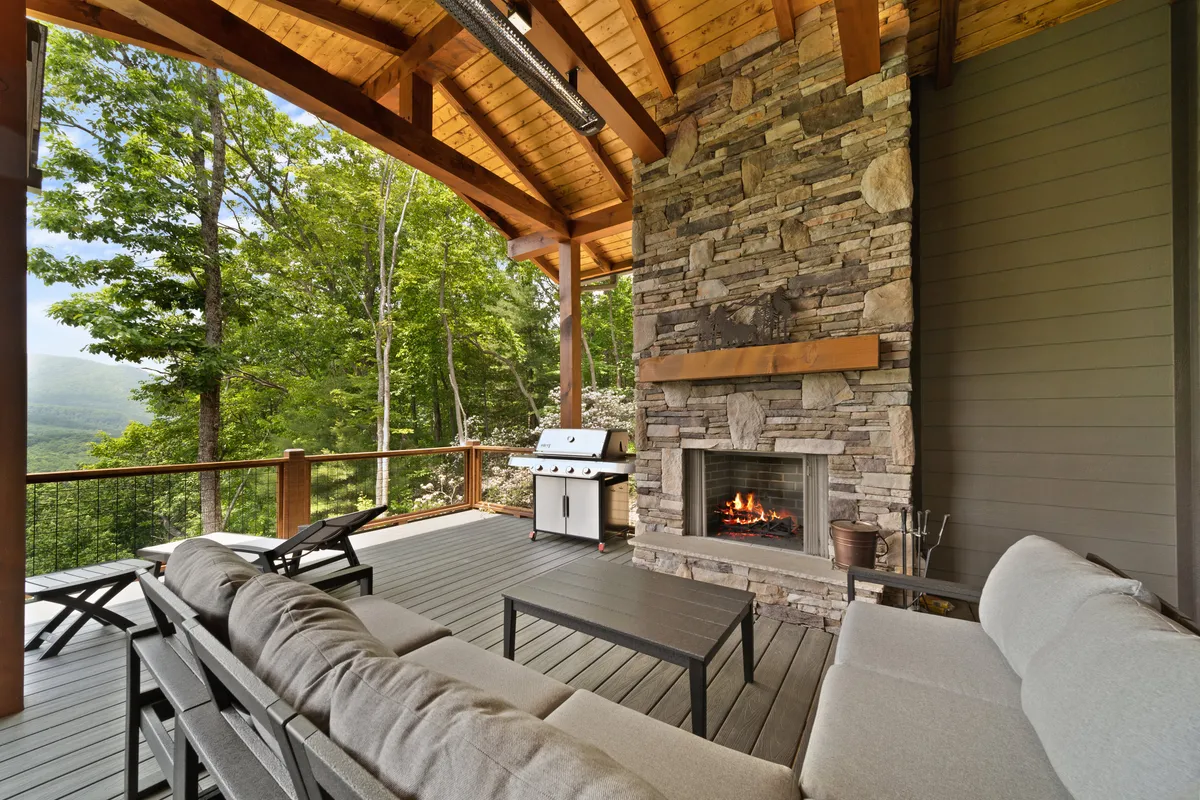 A cozy exterior living area centered around a stone outdoor fireplace with comfortable seating and a grilling area. The mountain views and sunset ambiance enhance the inviting atmosphere, signaling warmth and social connection for gatherings or quiet evenings by the fire.