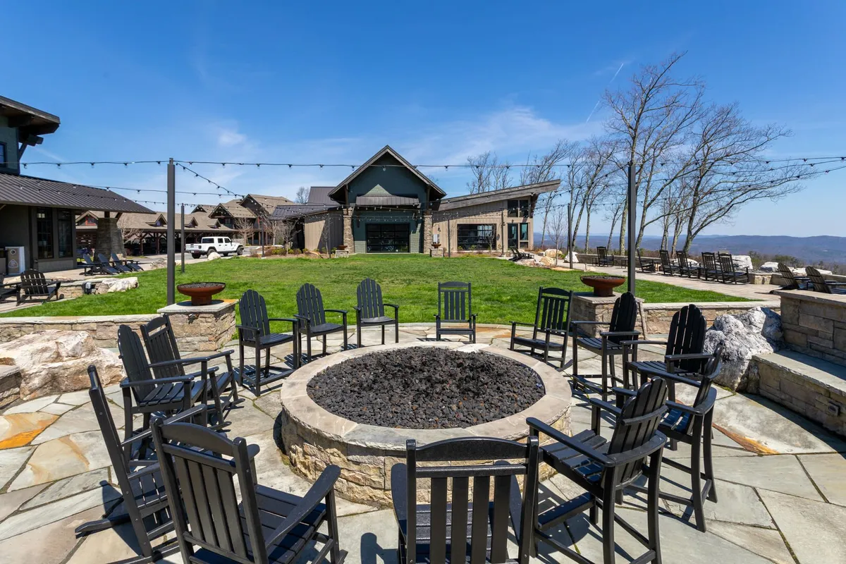 A cozy fire pit surrounded by wooden chairs on a landscaped lawn, overlooking distant mountain views. The scene suggests evenings spent gathering, sharing stories, and enjoying the natural setting as the sun sets or stars emerge. The vibe is inviting, communal, and quintessentially mountain-club.