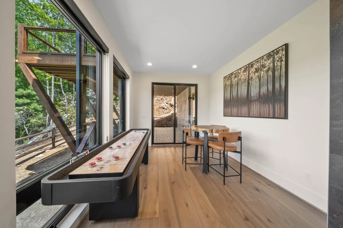 A shuffleboard table is the focal point of this inviting space, set near large windows that overlook a forested view. The image also shows a dining table and wall art, enhancing the sense of a relaxed, multi-use recreation area.