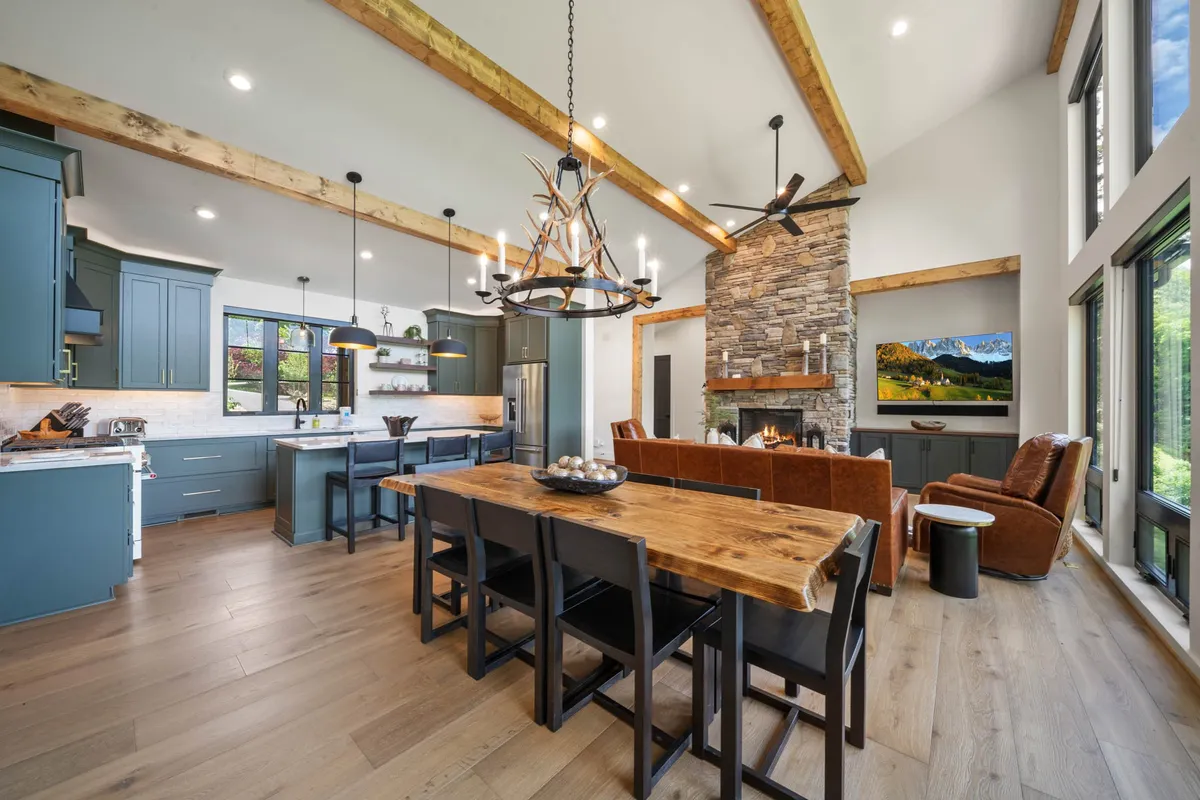 An open-concept living space with a stone fireplace, a large dining table set for gatherings, and a modern kitchen in the background. The comfortable seating, elegant chandelier, and kitchen amenities suggest a place ideal for both relaxing and hosting festive meals. The fireplace adds warmth and a touch of luxury mountain living.