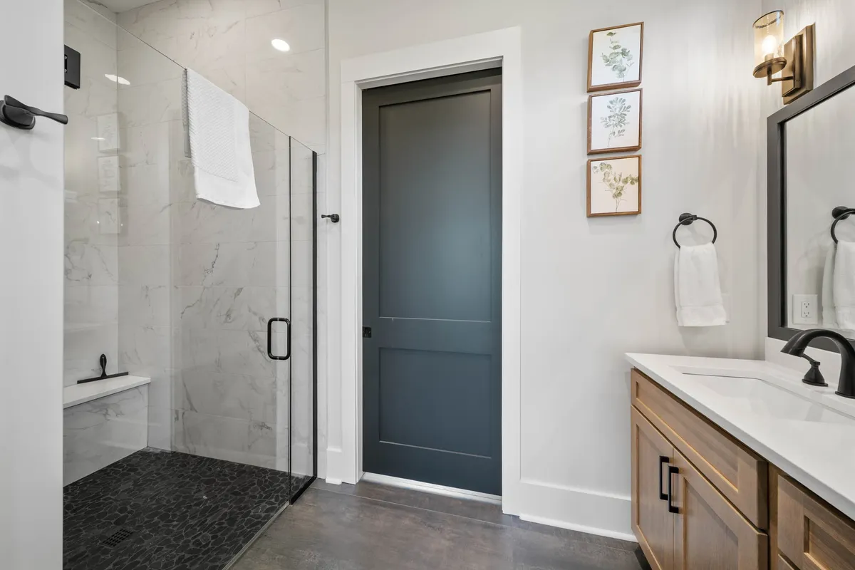 A bright, modern bathroom with a glass shower enclosure, dark door, wooden vanity, and framed botanical prints. The space signals luxury and serves as an ideal ensuite for the main-level king suite.