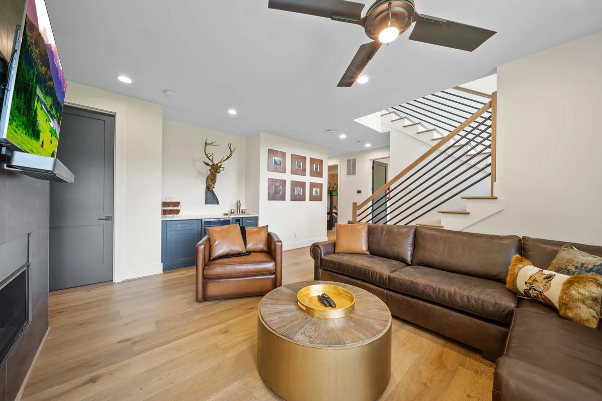 This cozy secondary living area includes a leather sofa, wooden coffee table, TV mounted on the wall, decorative wall art, and a visible staircase. The layout is inviting for groups to gather, watch movies, or relax together after a day of activities.