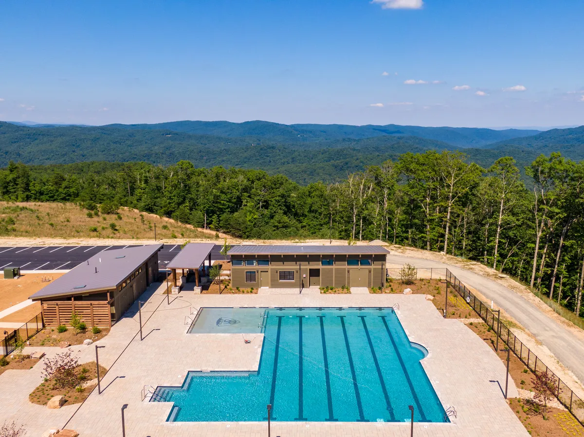 This image showcases a large, sparkling outdoor swimming pool set against a breathtaking mountain landscape. A modern pool house sits nearby, with the entire scene bathed in natural light and surrounded by rolling hills and greenery, creating a sense of both fun and tranquility. The setting hints at community social gatherings and family-friendly relaxation.