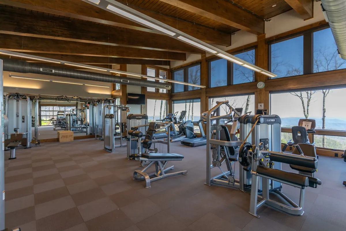 A modern fitness center with exercise machines set against large windows, allowing natural light to stream in and offering views of the outdoors. Wooden beams and natural materials create a warm, motivating environment. The room is versatile, suitable for yoga, group fitness, and personal training.