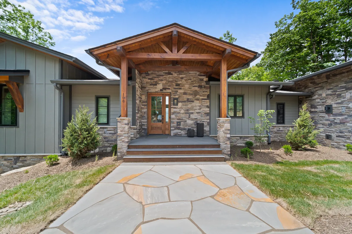 The image showcases the front entrance of a beautiful single-family home, featuring a striking stone facade, elegant wooden beams, and a lush, meticulously landscaped garden. The inviting porch hints at cozy evening gatherings, all set against the scenic backdrop of the Blue Ridge Mountains.